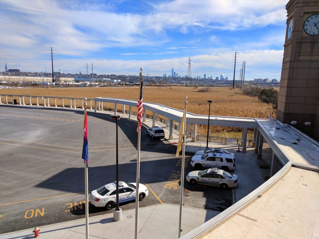 This image shows an outdoor parking area during the day, with a clear blue sky above. There are three transit police vehicles parked in the lot, which is otherwise empty. A sprawling brown field lies beyond the parking area, leading to a distant city skyline under a hazy sky.