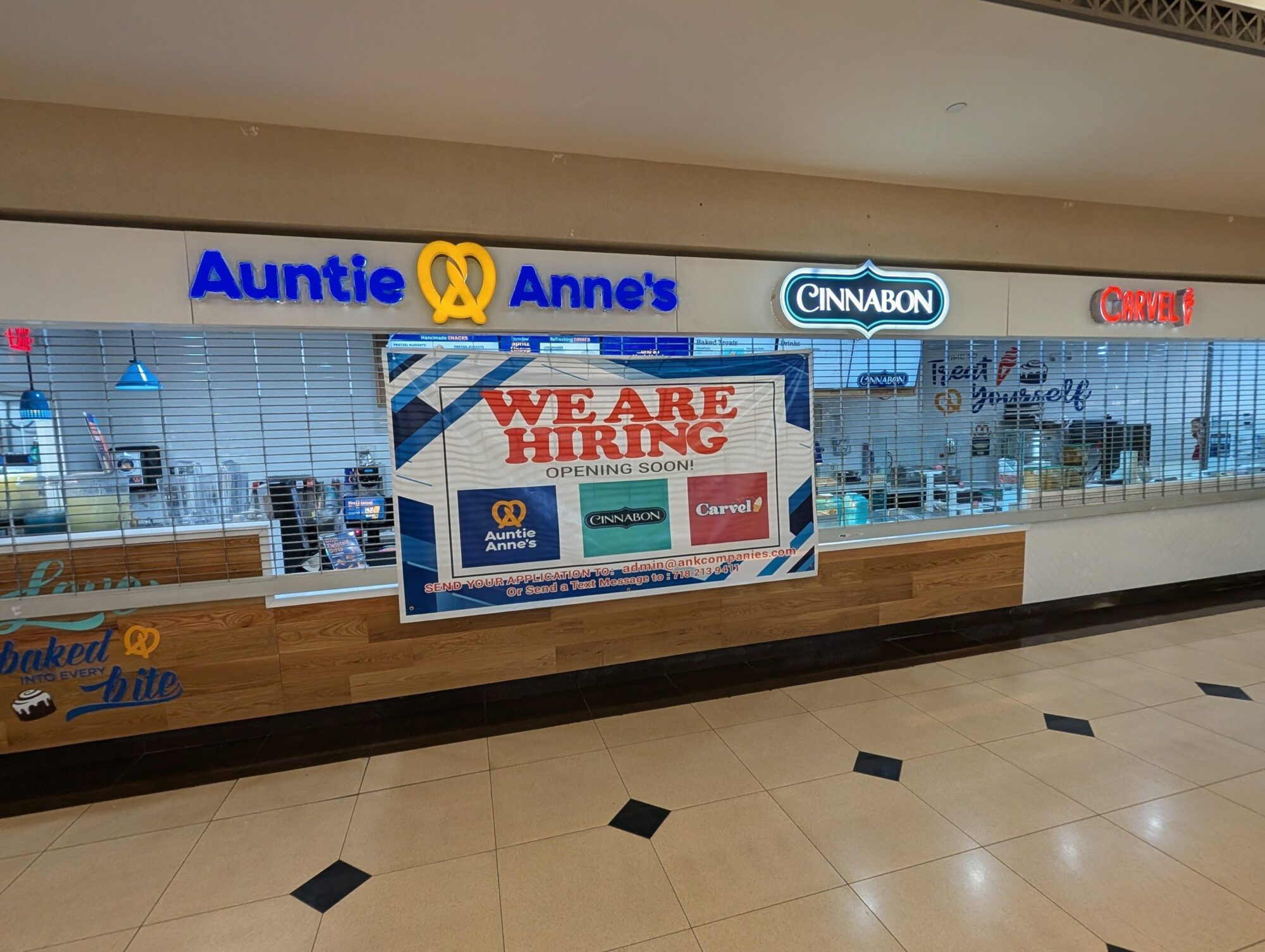 A brightly lit, newly installed storefront sign inside a train station concourse. The sign features the logos for Auntie Anne's, Cinnabon, and Carvel. A large "WE ARE HIRING" banner is centered in the window, with the store's metal security gates partially lowered.